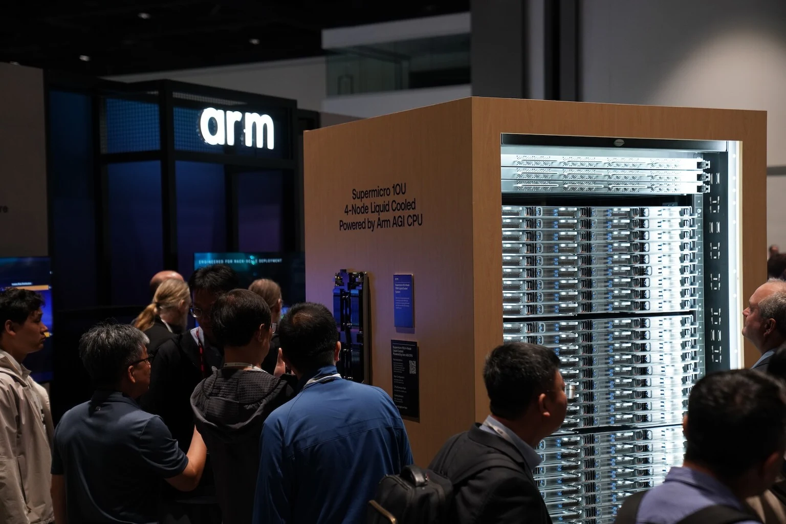 A liquid-cooled server rack is displayed at an Arm booth, with attendees gathered around it.