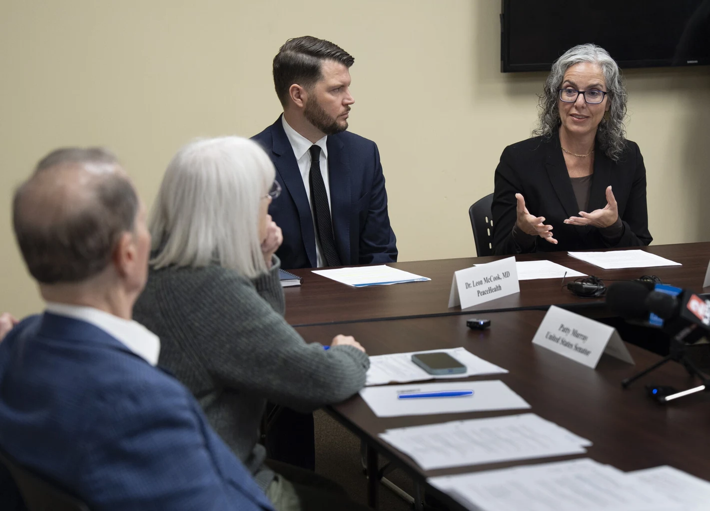 A woman speaks at a meeting table while others listen; placards for Dr. Leon McCook and Patty Murray are visible.